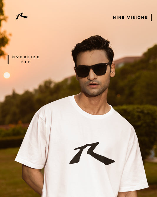 Man wearing sunglasses and a white t-shirt with a logo, standing outdoors with trees and sunset in the background.